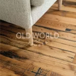 Close-up of reclaimed industrial oak floor beneath a beige fabric armchair, displaying dark character streaks and wear patterns from historic use.